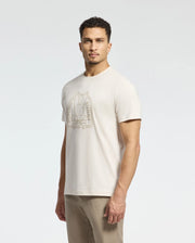 The man wears a Psycho Bunny MENS MELBOURNE GRAPHIC TEE - B6U410F200 made of eco-friendly Peruvian Pima cotton with a raised abstract animal design on the chest. He stands against a plain white background, looking towards the camera, paired with matching light beige pants.