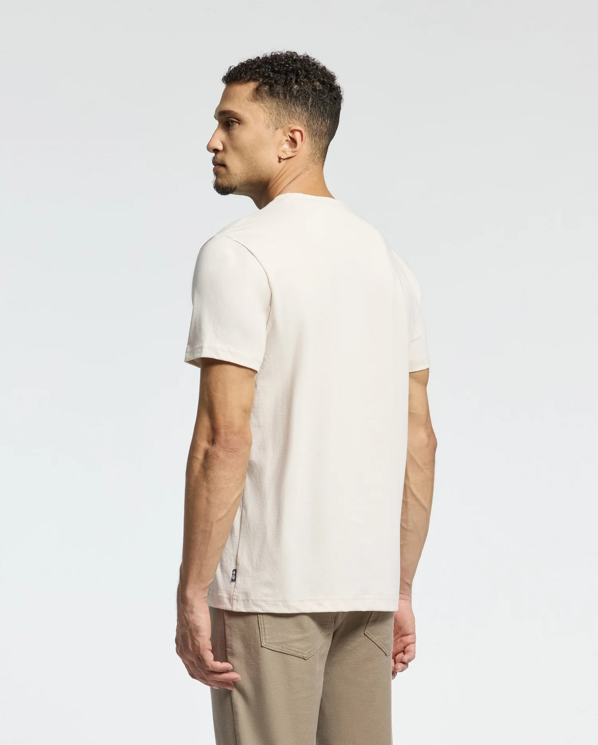 A man with short curly hair wears a cream-colored Psycho Bunny MENS MELBOURNE GRAPHIC TEE made of eco-friendly Peruvian Pima cotton and beige pants, standing partially visible against a plain, light gray background.