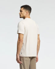 A man with short curly hair wears a cream-colored Psycho Bunny MENS MELBOURNE GRAPHIC TEE made of eco-friendly Peruvian Pima cotton and beige pants, standing partially visible against a plain, light gray background.