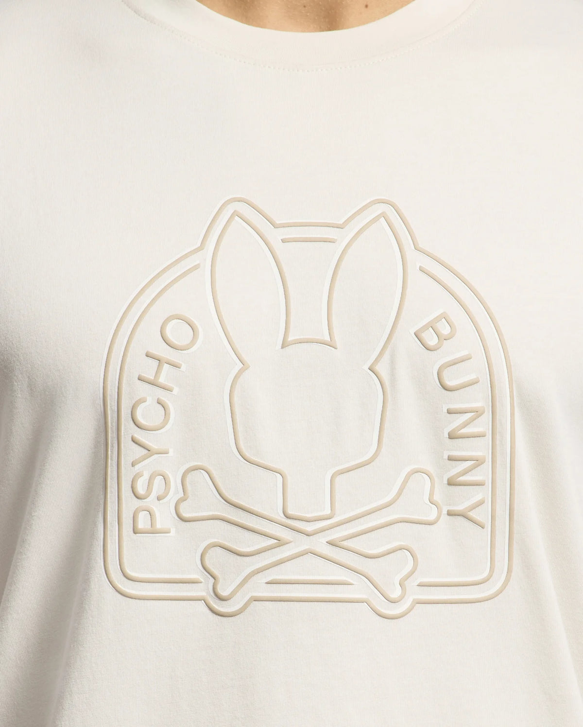 Close-up of a cream-colored Psycho Bunny Men's Melbourne Graphic Tee, made from eco-friendly Peruvian Pima cotton, featuring an embossed stylized bunny head and crossbones with "Psycho Bunny" embossed vertically on either side.