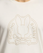 Close-up of a cream-colored Psycho Bunny Men's Melbourne Graphic Tee, made from eco-friendly Peruvian Pima cotton, featuring an embossed stylized bunny head and crossbones with "Psycho Bunny" embossed vertically on either side.