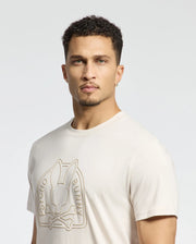 A person with short curly hair and a goatee is wearing the eco-friendly Psycho Bunny MENS MELBOURNE GRAPHIC TEE - B6U410F200, made of luxurious Peruvian Pima cotton, featuring a rabbit above crossed bones graphic. The background is plain white or light gray.
