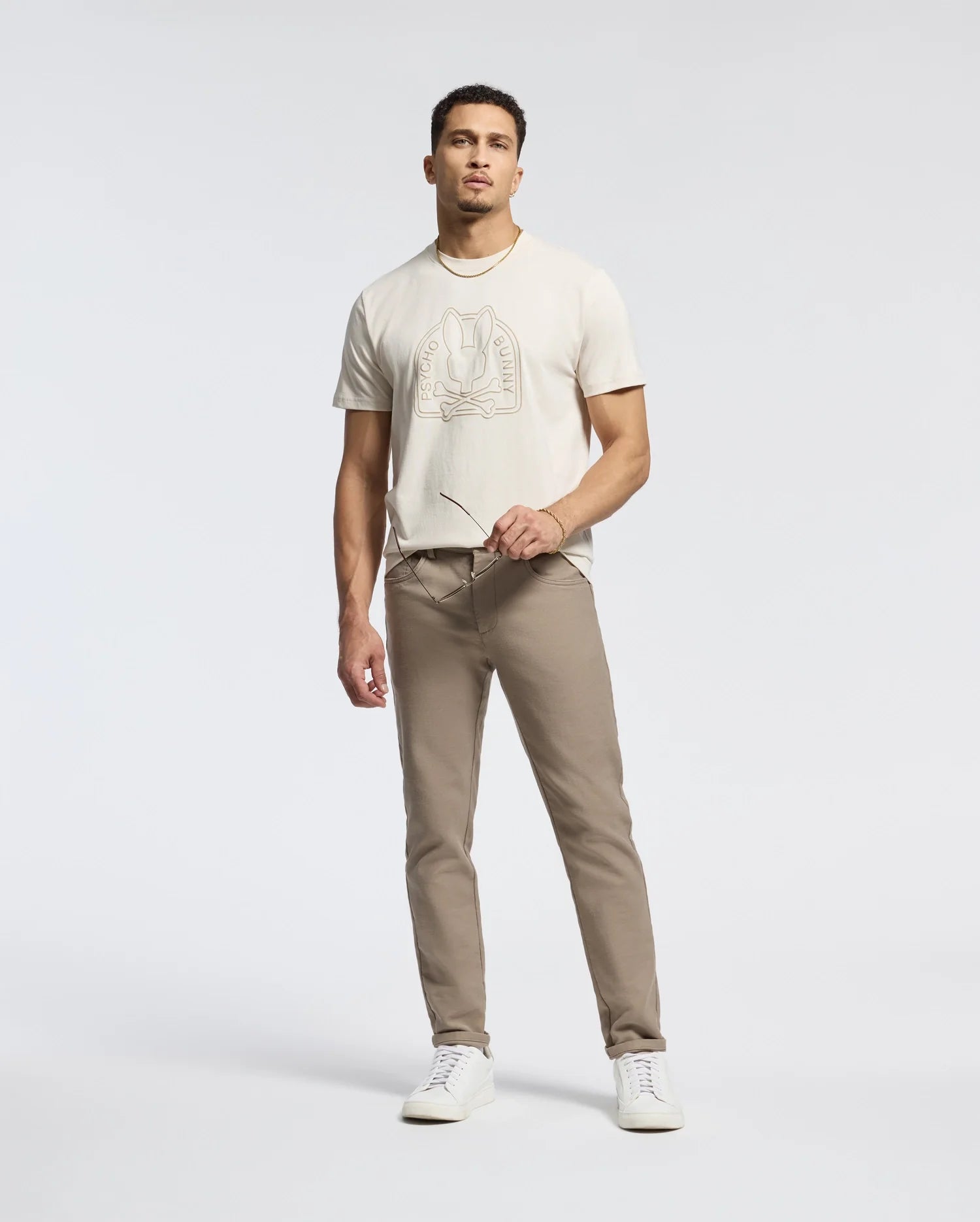 A man stands against a plain background wearing a light-colored Psycho Bunny MENS MELBOURNE GRAPHIC TEE - B6U410F200 made from eco-friendly Peruvian Pima cotton, beige pants, and white sneakers. He holds a pair of glasses in one hand and gazes forward with a neutral expression.