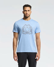 A man with short curly hair and a beard models the eco-friendly, light blue Psycho Bunny MENS MELBOURNE GRAPHIC TEE - B6U410F200 featuring a bunny graphic with crossbones and "Psycho Bunny" text. He stands against a white background, paired with black pants.