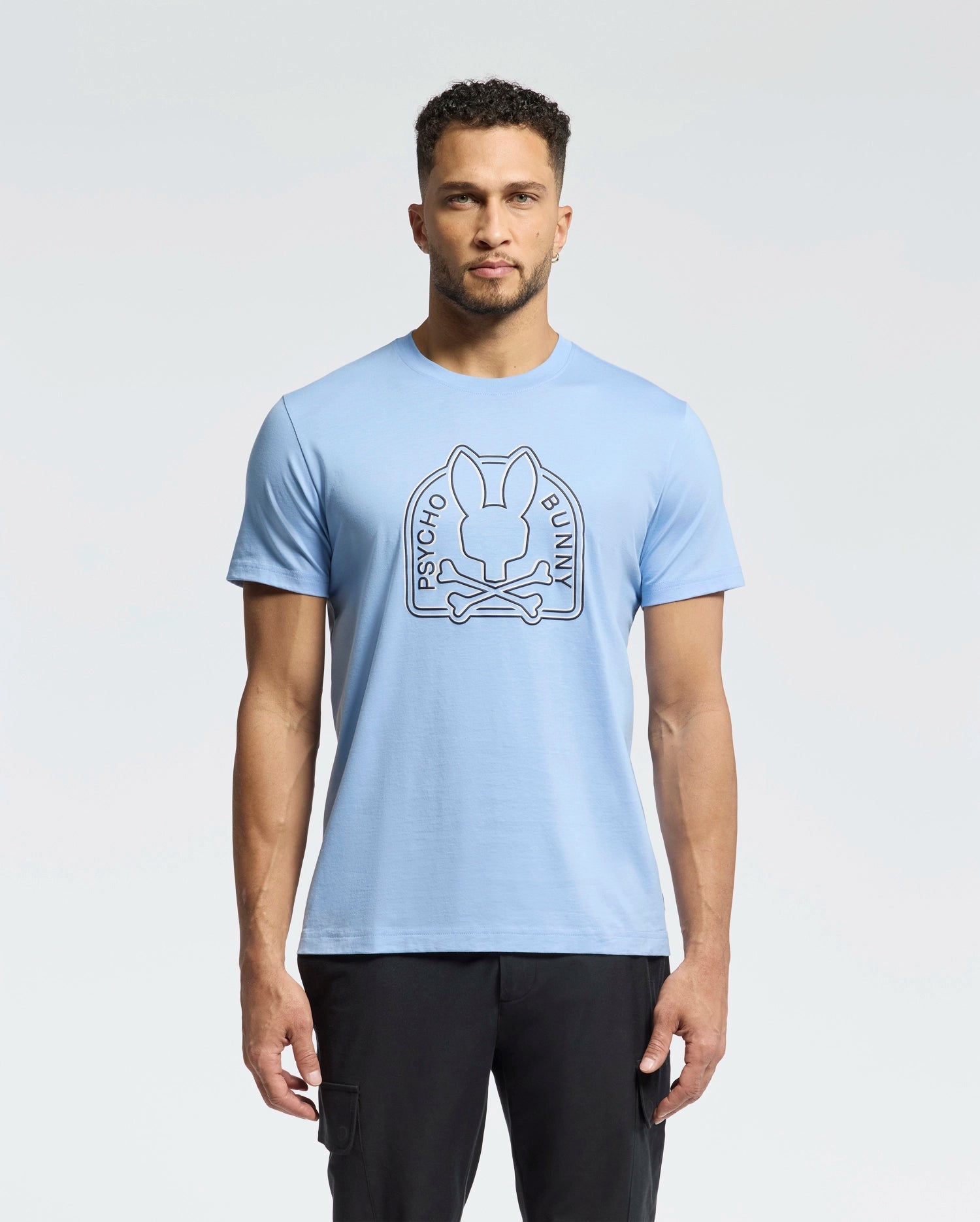 A man with short curly hair and a beard models the eco-friendly, light blue Psycho Bunny MENS MELBOURNE GRAPHIC TEE - B6U410F200 featuring a bunny graphic with crossbones and "Psycho Bunny" text. He stands against a white background, paired with black pants.