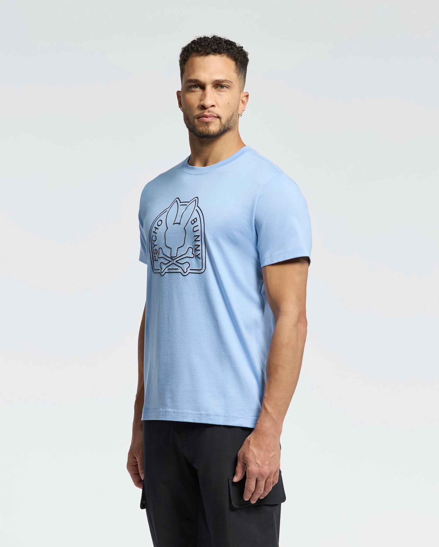 A person with short curly hair wears a light blue Men's Melbourne Graphic Tee (B6U410F200) by Psycho Bunny featuring a stylized rabbit. They stand against a plain background, looking directly at the camera, blending style and sustainability.