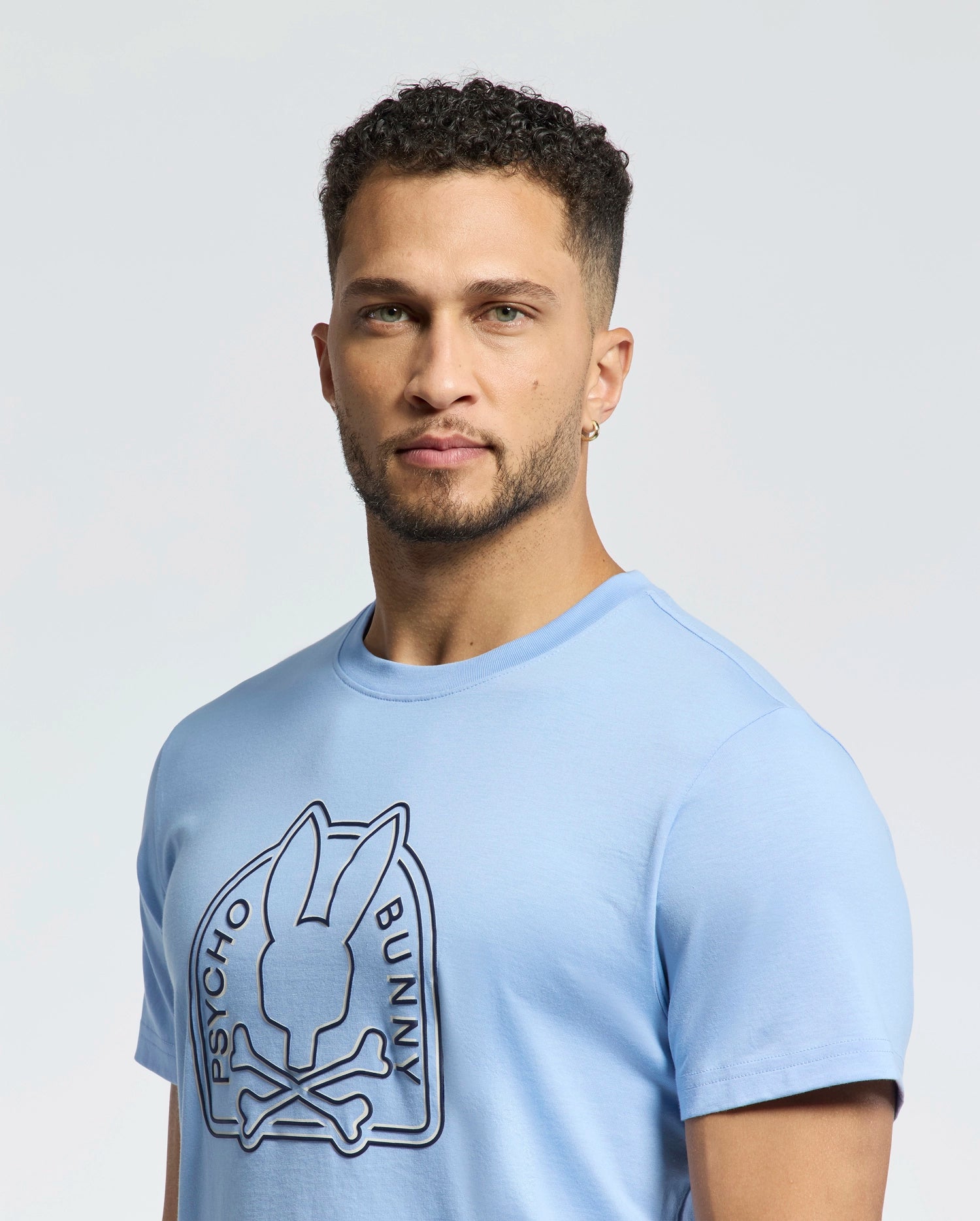 Someone wearing a Psycho Bunny MENS MELBOURNE GRAPHIC TEE (B6U410F200) in light blue stands against a plain background. Made from eco-friendly Peruvian Pima cotton, the tee features a stylized bunny and crossbones design in dark print.