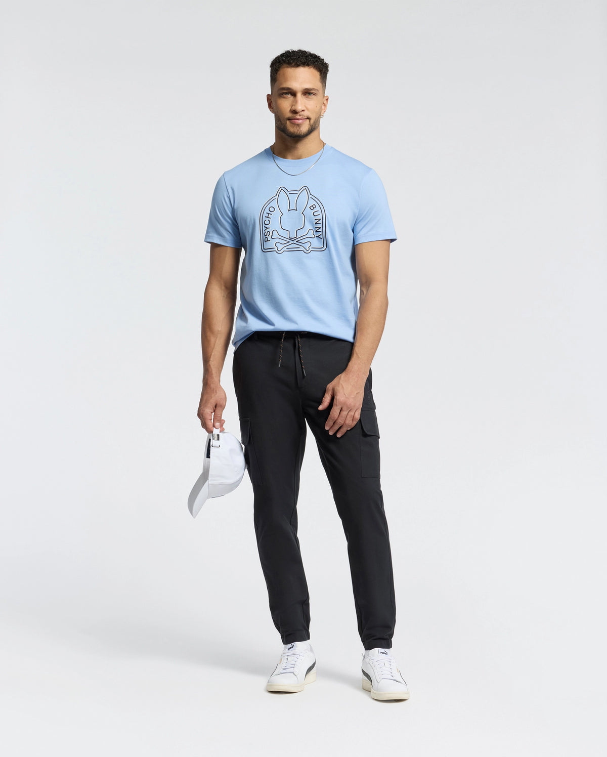 A man confidently stands against a plain background, wearing a Psycho Bunny Peruvian Pima Melbourne tee in light blue with a graphic design, paired with black joggers and white sneakers. He holds an eco-friendly white cap in his left hand.