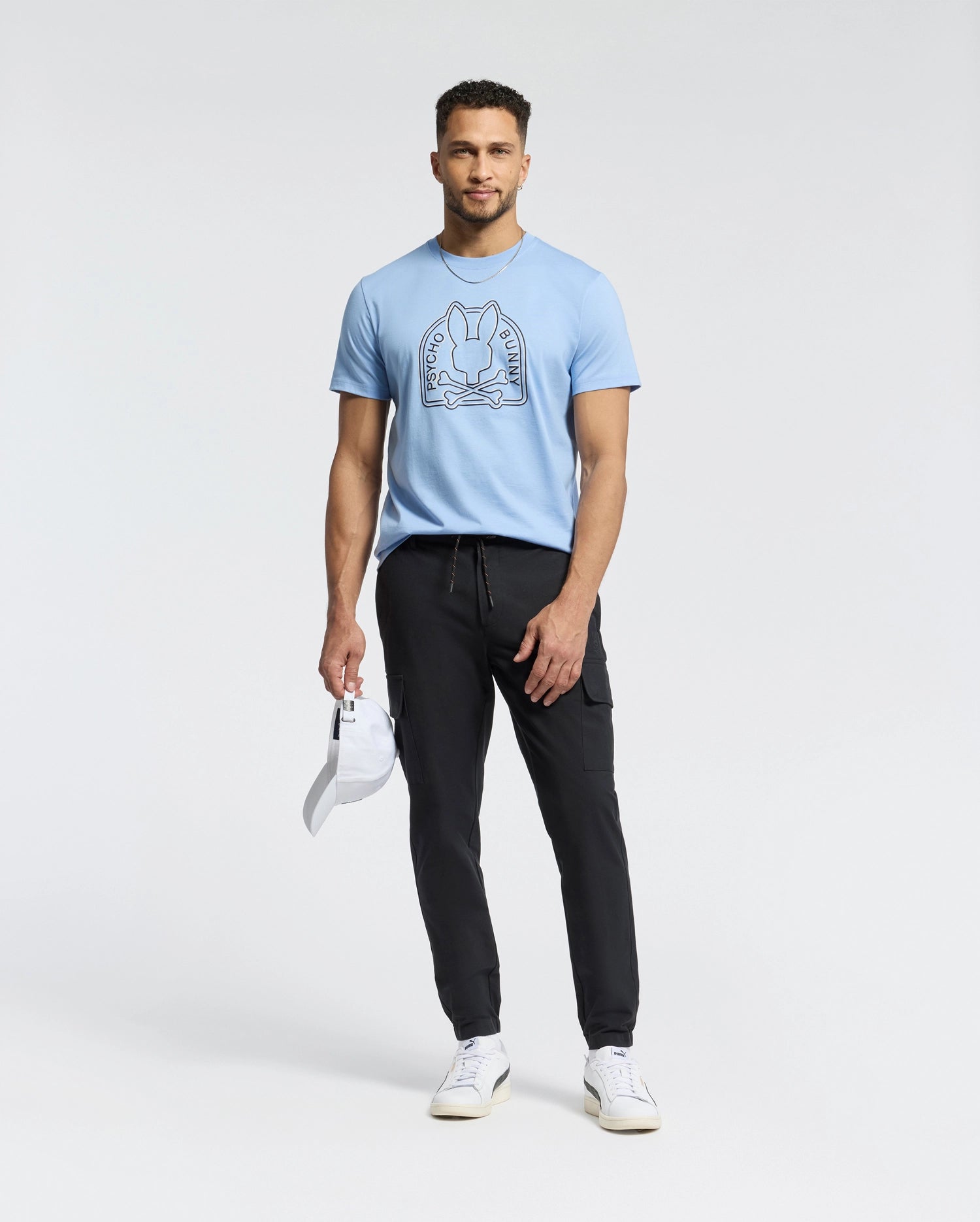 A man confidently stands against a plain background, wearing a Psycho Bunny Peruvian Pima Melbourne tee in light blue with a graphic design, paired with black joggers and white sneakers. He holds an eco-friendly white cap in his left hand.