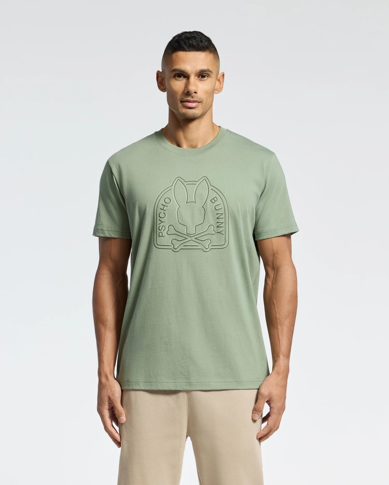 A man stands facing forward in a light green Psycho Bunny MENS MELBOURNE GRAPHIC TEE (B6U410F200) with a large embossed bunny and crossbones logo, paired with beige pants, against a plain light background.