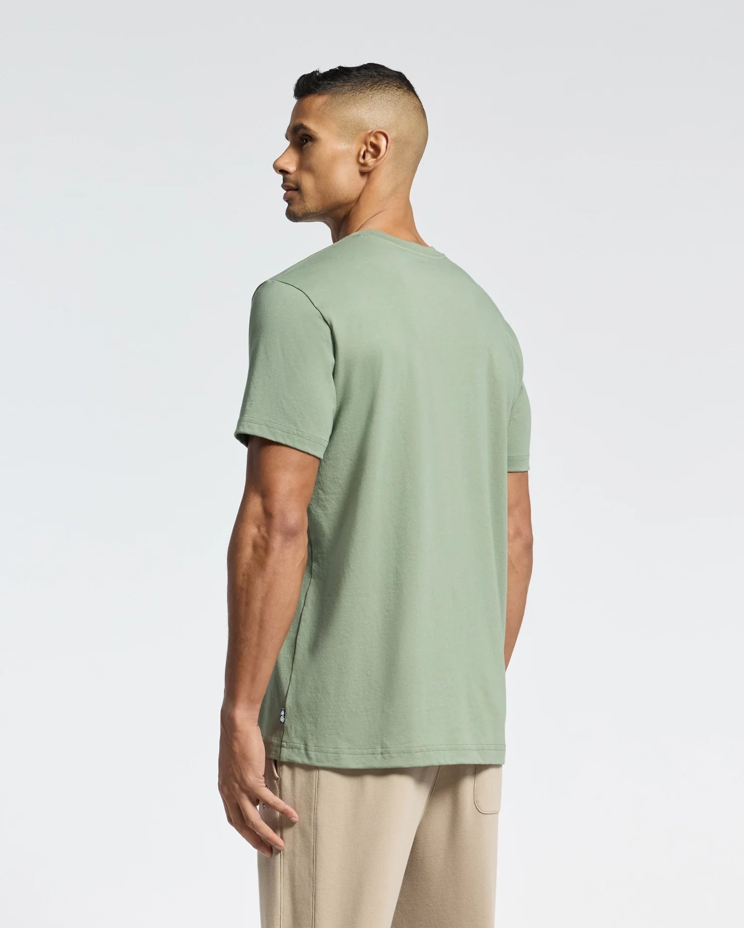 A man with short dark hair stands facing away, wearing the Psycho Bunny MENS MELBOURNE GRAPHIC TEE - B6U410F200 in light green and beige pants against a plain, light background.
