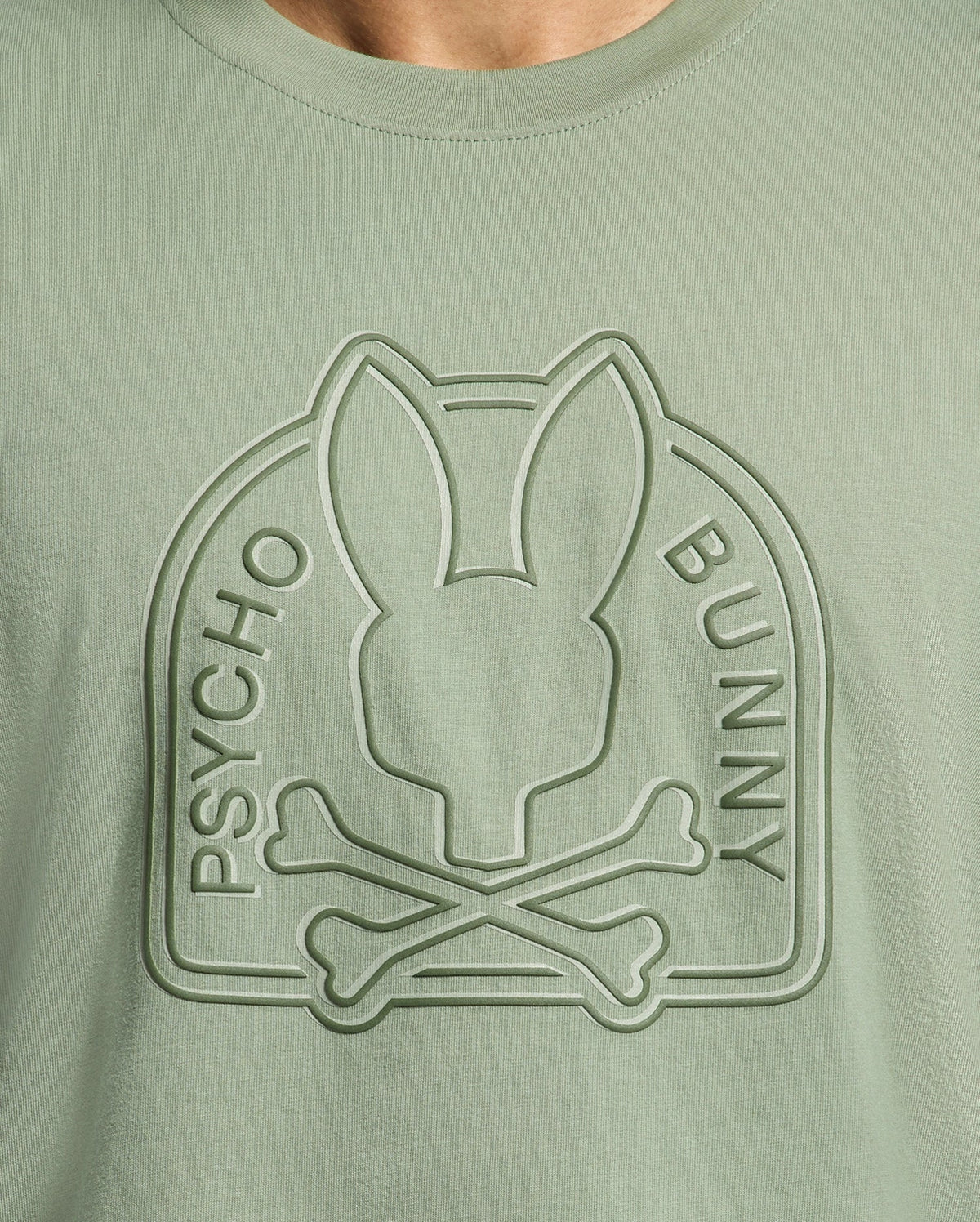 Close-up of the light green Psycho Bunny MENS MELBOURNE GRAPHIC TEE (B6U410F200), featuring an eco-friendly embossed bunny head above crossed bones, an arch frame, and "PSYCHO BUNNY" on either side of the logo.