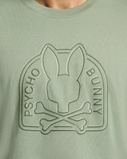 Close-up of the light green Psycho Bunny MENS MELBOURNE GRAPHIC TEE (B6U410F200), featuring an eco-friendly embossed bunny head above crossed bones, an arch frame, and "PSYCHO BUNNY" on either side of the logo.