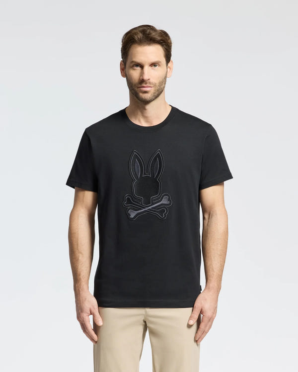 A man wearing Psycho Bunny's MENS NEW CASTLE EMBROIDERED GRAPHIC TEE, featuring a bunny skull and crossbones, stands against a white background. With short hair, he pairs the tee with khaki pants.