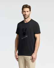 A short-haired, bearded man wears a Psycho Bunny "MENS NEW CASTLE EMBROIDERED GRAPHIC TEE - B6U415F200" in black Pima cotton with an embroidered bunny skull design, paired with beige pants against a plain light gray backdrop.