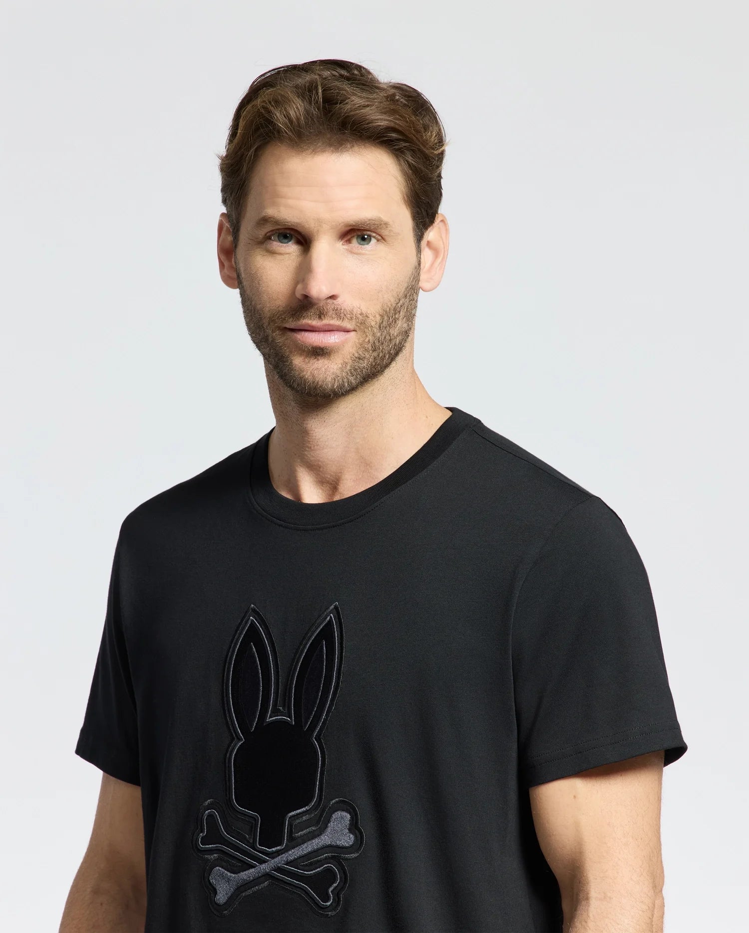 A man with short brown hair and a beard wears the Psycho Bunny MENS NEW CASTLE EMBROIDERED GRAPHIC TEE, showcasing a bunny head above crossbones on a black Pima cotton fabric, set against a plain gray background.