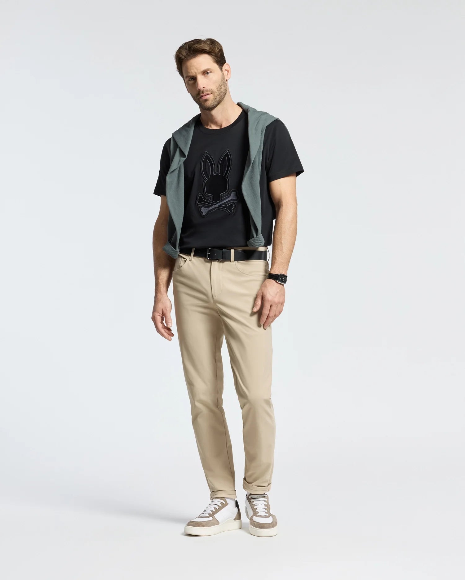 A man wears a Psycho Bunny New Castle embroidered graphic tee in black, beige pants, and sneakers. He has a green jacket draped over his shoulders with a watch and belt. The background is plain white.