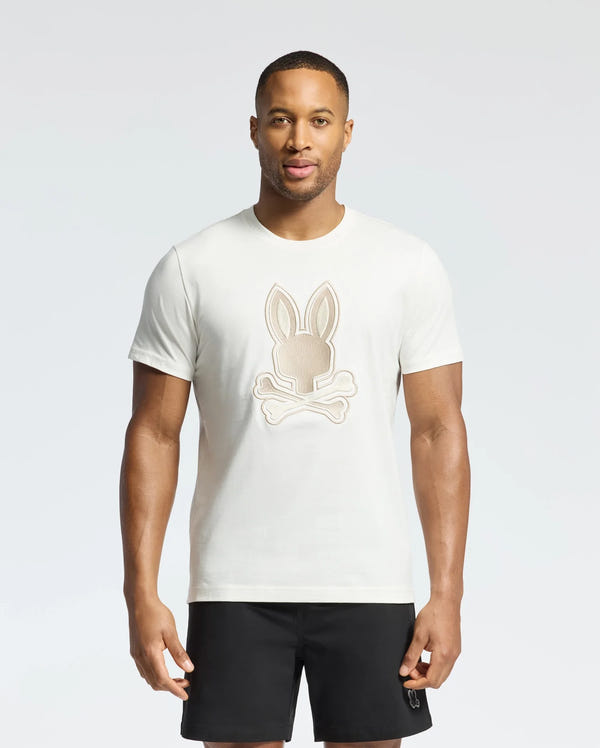 A person is wearing a Psycho Bunny MENS NEW CASTLE EMBROIDERED GRAPHIC TEE in white, featuring an embroidered Bunny and crossbones design on the front, paired with black shorts against a plain white background.