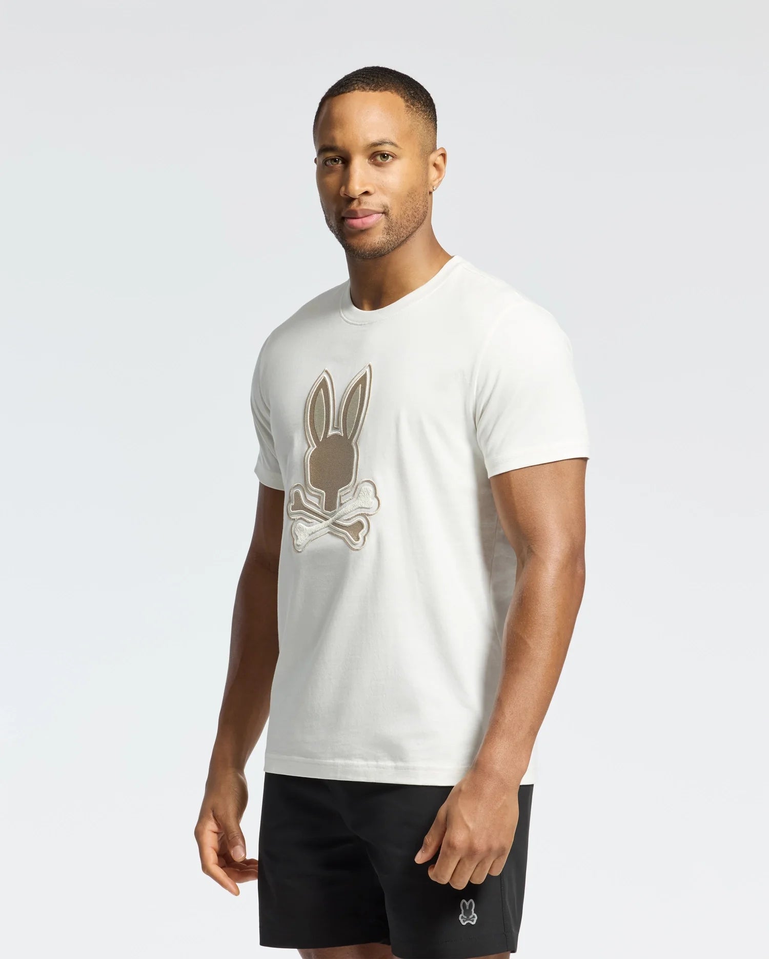 A man confidently stands against a white background, wearing the Psycho Bunny MENS NEW CASTLE EMBROIDERED GRAPHIC TEE and black shorts, looking directly at the camera.