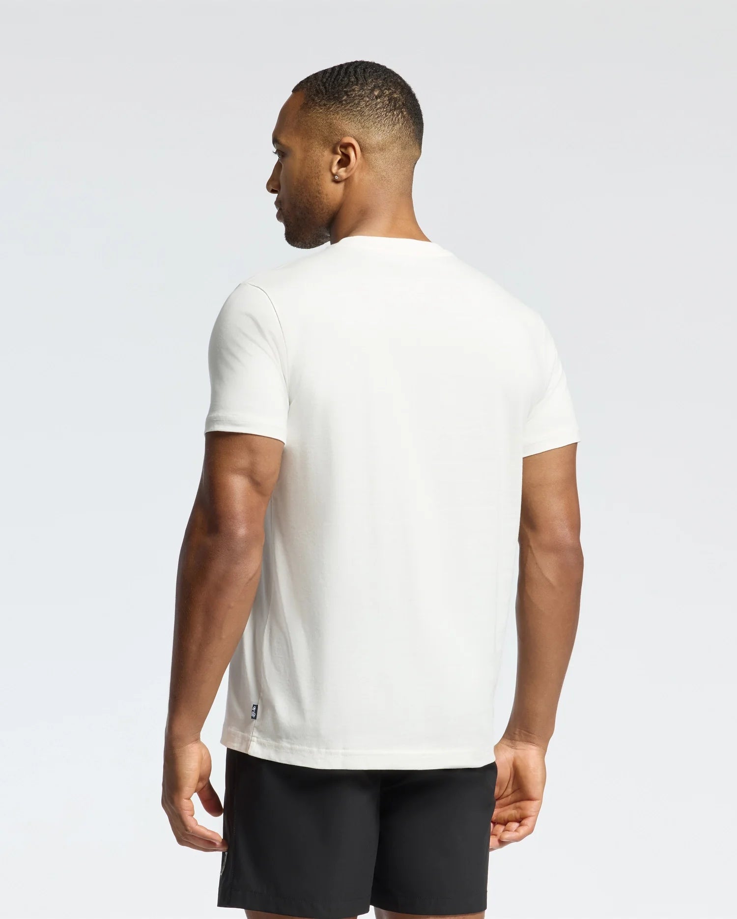 A person wearing a Psycho Bunny MEN'S NEW CASTLE EMBROIDERED GRAPHIC TEE, made from Pima cotton, and black shorts is standing and facing away from the camera against a light gray background.