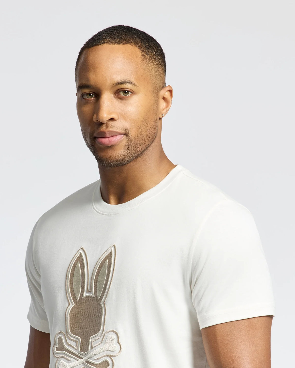 A short-haired person with an earring wears Psycho Bunny's MENS NEW CASTLE EMBROIDERED GRAPHIC TEE (B6U415F200), a white Pima cotton shirt featuring a playful bunny with crossbones, set against a plain, light-colored background.
