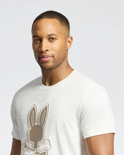A short-haired person with an earring wears Psycho Bunny's MENS NEW CASTLE EMBROIDERED GRAPHIC TEE (B6U415F200), a white Pima cotton shirt featuring a playful bunny with crossbones, set against a plain, light-colored background.