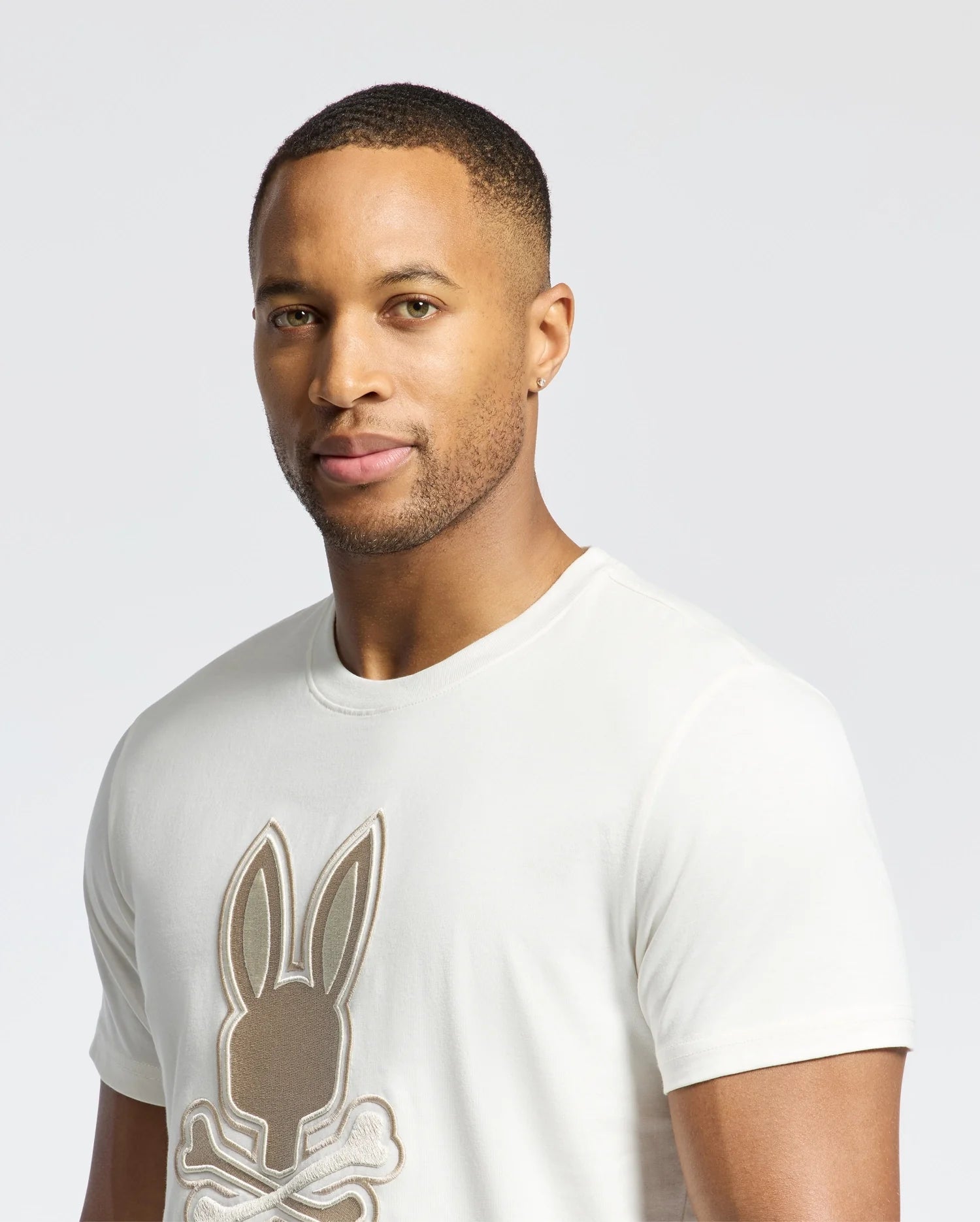 A short-haired person with an earring wears Psycho Bunny's MENS NEW CASTLE EMBROIDERED GRAPHIC TEE (B6U415F200), a white Pima cotton shirt featuring a playful bunny with crossbones, set against a plain, light-colored background.