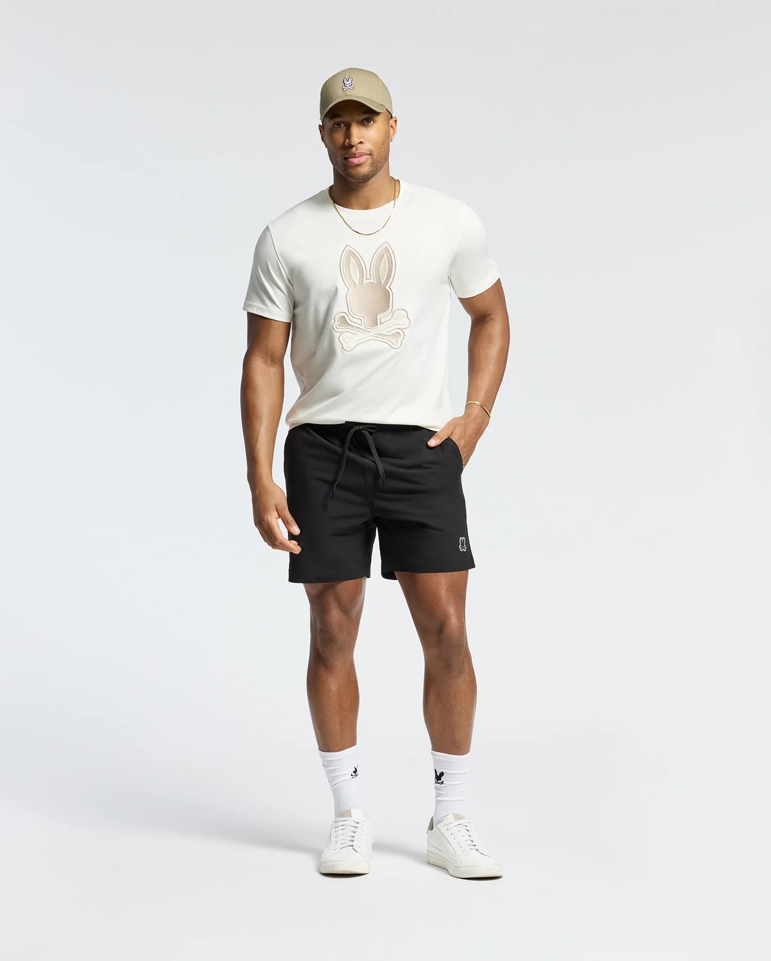 A person in a beige cap and a Psycho Bunny MENS NEW CASTLE EMBROIDERED GRAPHIC TEE featuring a bunny design poses against a neutral backdrop. They wear black shorts, white socks, and sneakers with their hands resting casually by their sides.