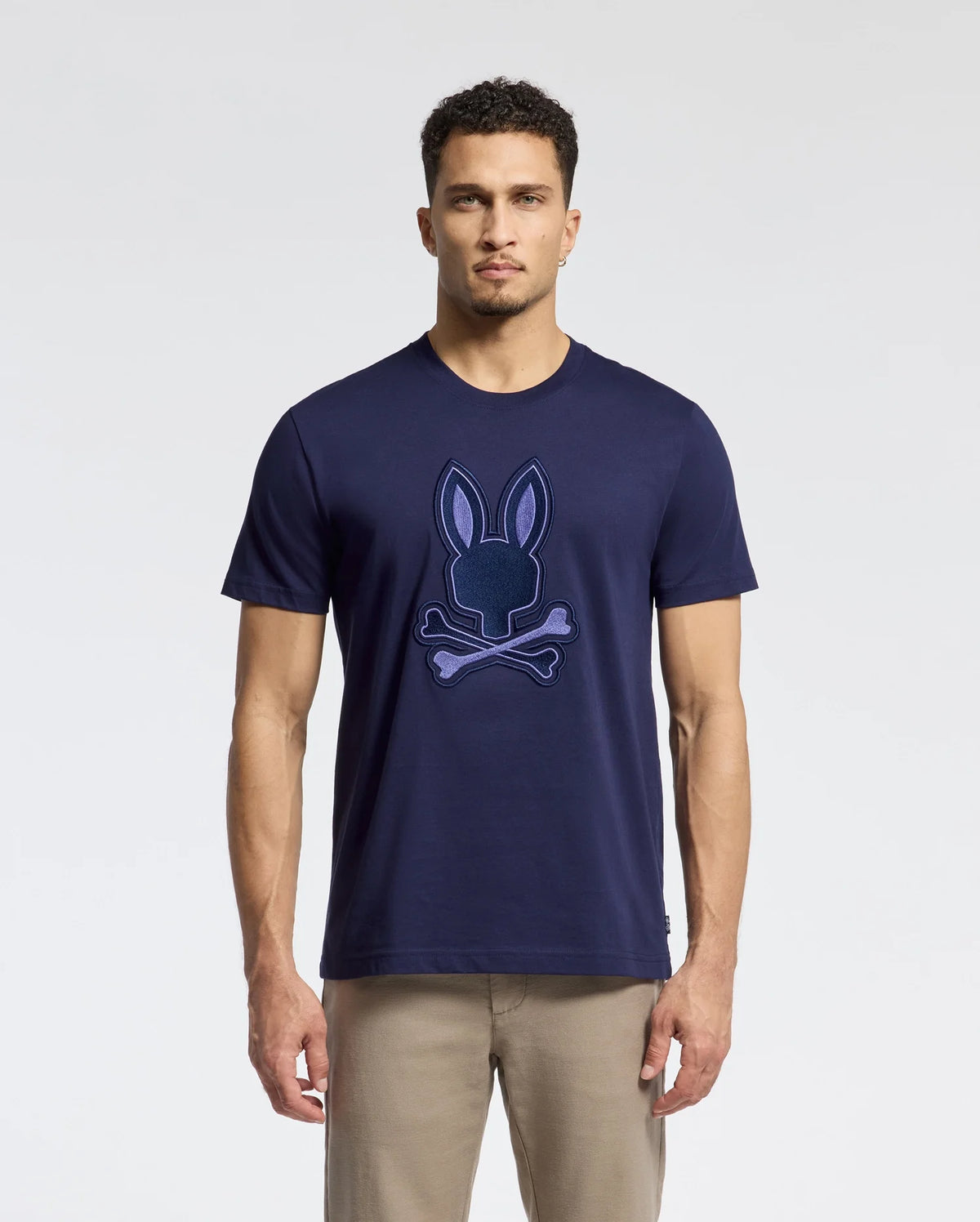 A man stands against a white background, wearing the Psycho Bunny Men's New Castle Embroidered Graphic Tee (B6U415F200) in navy blue, featuring an embroidered Bunny skull and crossbones logo on the front, paired with beige pants.