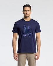 A man stands against a white background, wearing the Psycho Bunny Men's New Castle Embroidered Graphic Tee (B6U415F200) in navy blue, featuring an embroidered Bunny skull and crossbones logo on the front, paired with beige pants.