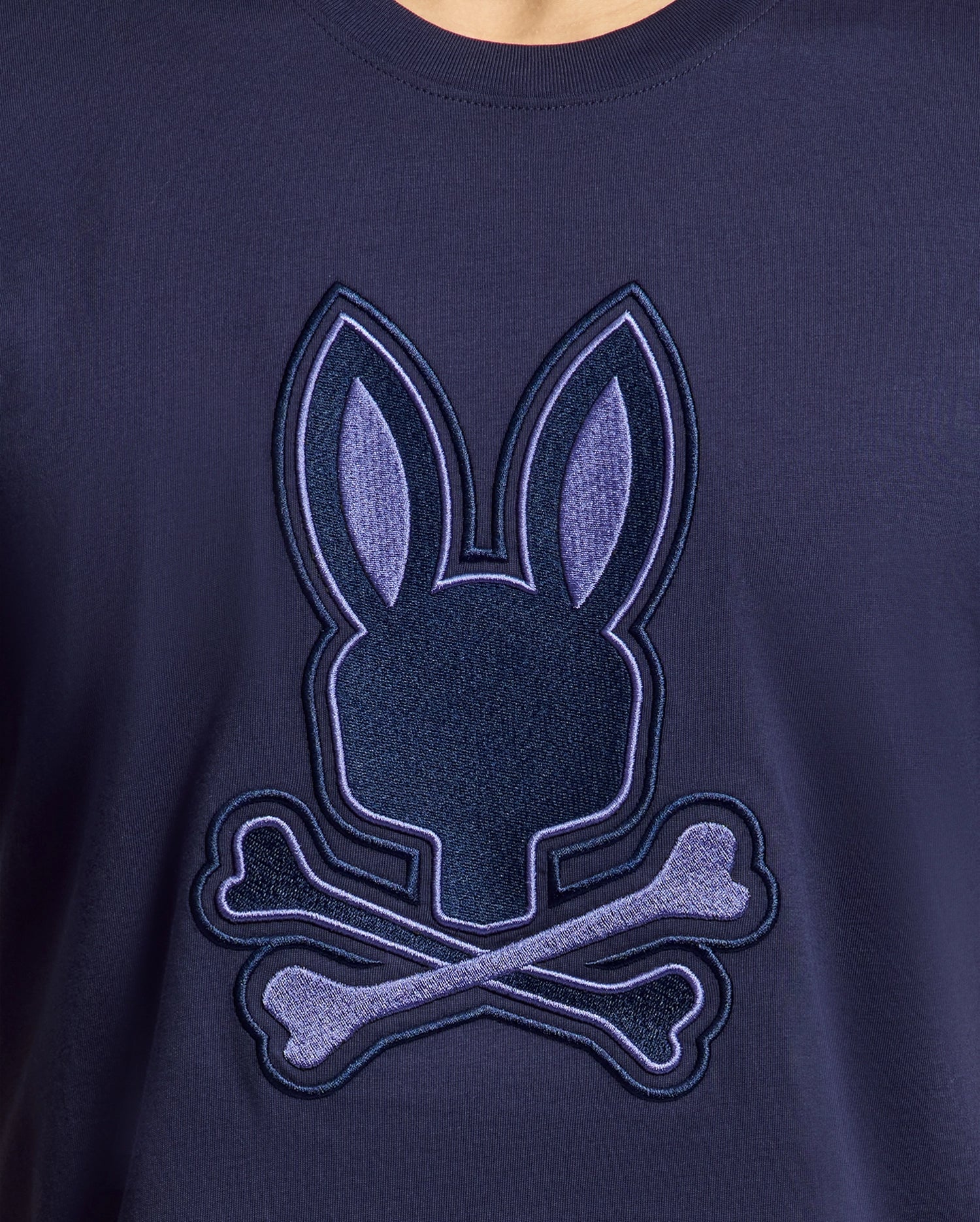 The Psycho Bunny MENS NEW CASTLE EMBROIDERED GRAPHIC TEE in navy blue features a soft Pima cotton fabric with an embroidered bunny head and crossed bones, using lighter blue shades for contrast.