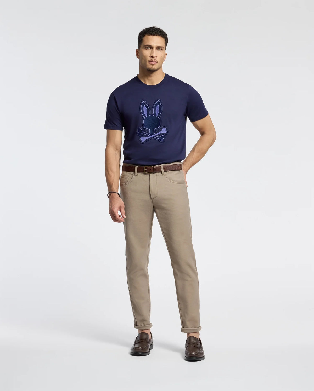 A man stands against a plain background, wearing the Psycho Bunny MENS NEW CASTLE EMBROIDERED GRAPHIC TEE in navy blue and khaki pants. He completes his look with brown shoes and a belt, maintaining a casual posture and neutral expression.