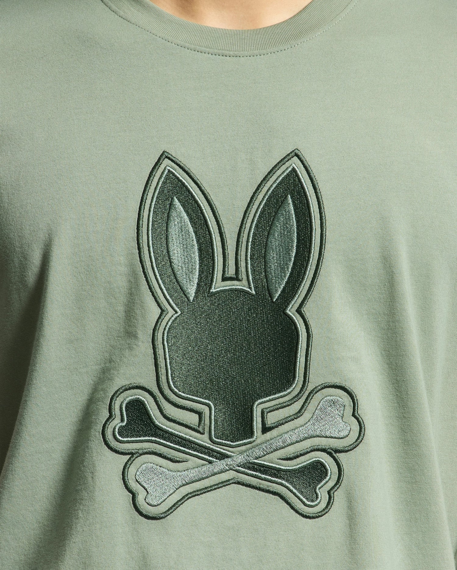 The MENS NEW CASTLE EMBROIDERED GRAPHIC TEE by Psycho Bunny features a playful twist on the classic skull and crossbones symbol with a large dark green stylized bunny head graphic above crossed bones, set on a soft light green Pima cotton fabric.