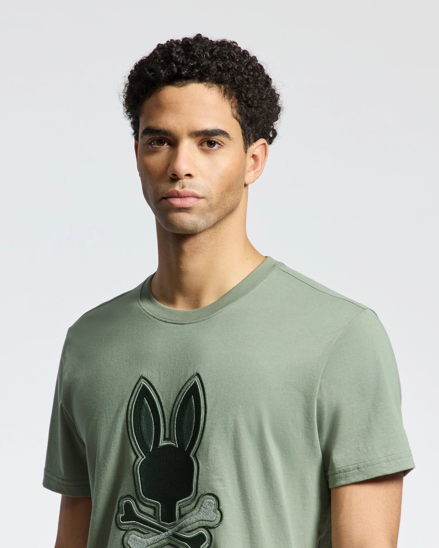 A man wearing the Psycho Bunny MENS NEW CASTLE EMBROIDERED GRAPHIC TEE - B6U415F200, featuring a black bunny and crossbones graphic, stands against a plain white background.