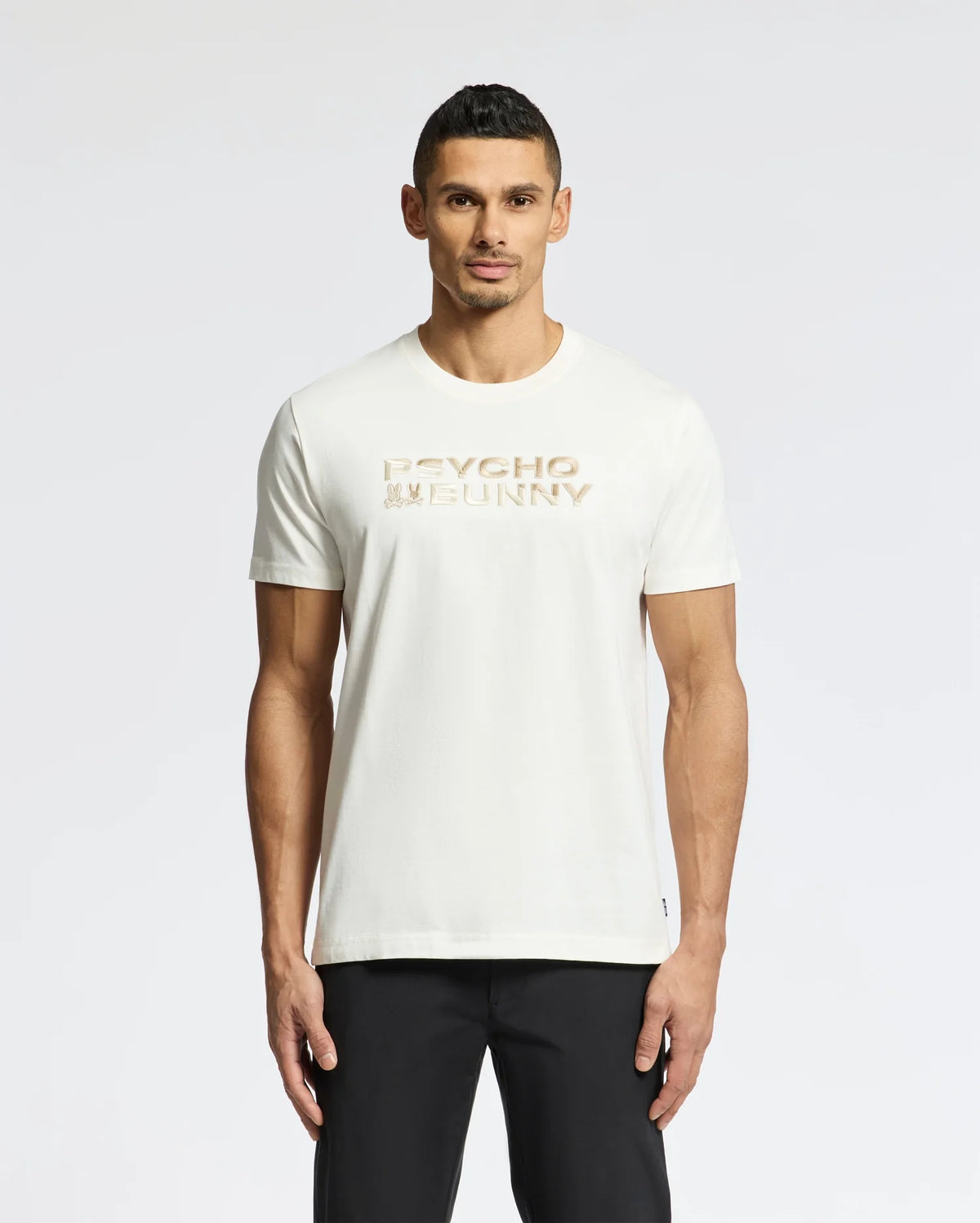 The man, with short dark hair and a neutral expression, stands against a plain white background wearing sleek black pants and the MENS BERNI EMBROIDERED WORDMARK GRAPHIC TEE - B6U417F200 by Psycho Bunny, featuring soft Pima cotton and a gold embroidered logo.