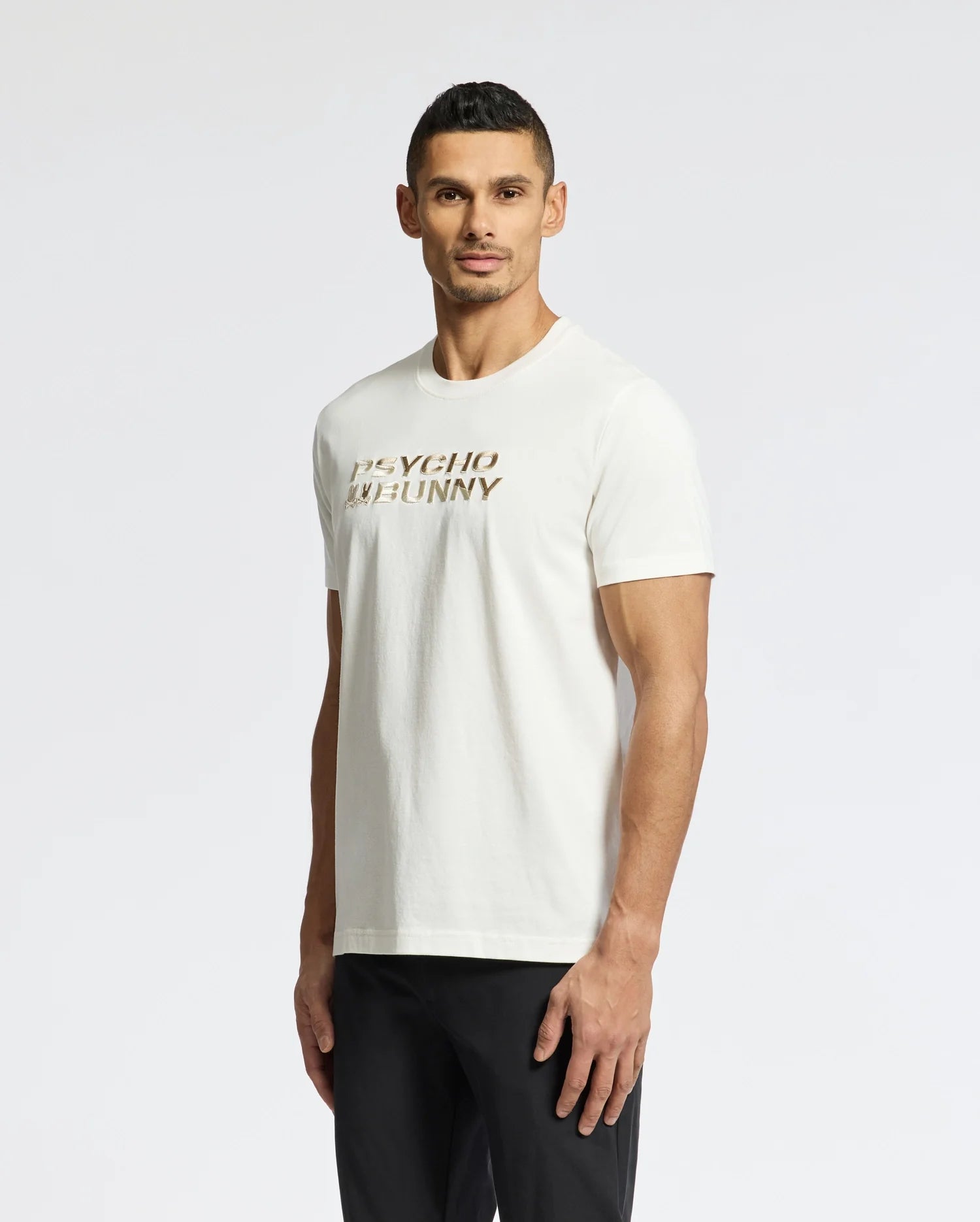 Facing forward, a person wears the MENS BERNI EMBROIDERED WORDMARK GRAPHIC TEE by Psycho Bunny in white, made from premium Pima cotton with "PSYCHO BUNNY" gradient print. They have short hair and black pants, set against a plain white background highlighting the embroidered logo.