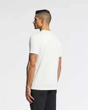 A man stands slightly sideways wearing a Psycho Bunny MENS BERNI EMBROIDERED WORDMARK GRAPHIC TEE - B6U417F200, crafted from soft Pima cotton with an embroidered logo, paired with black pants against a light gray background.