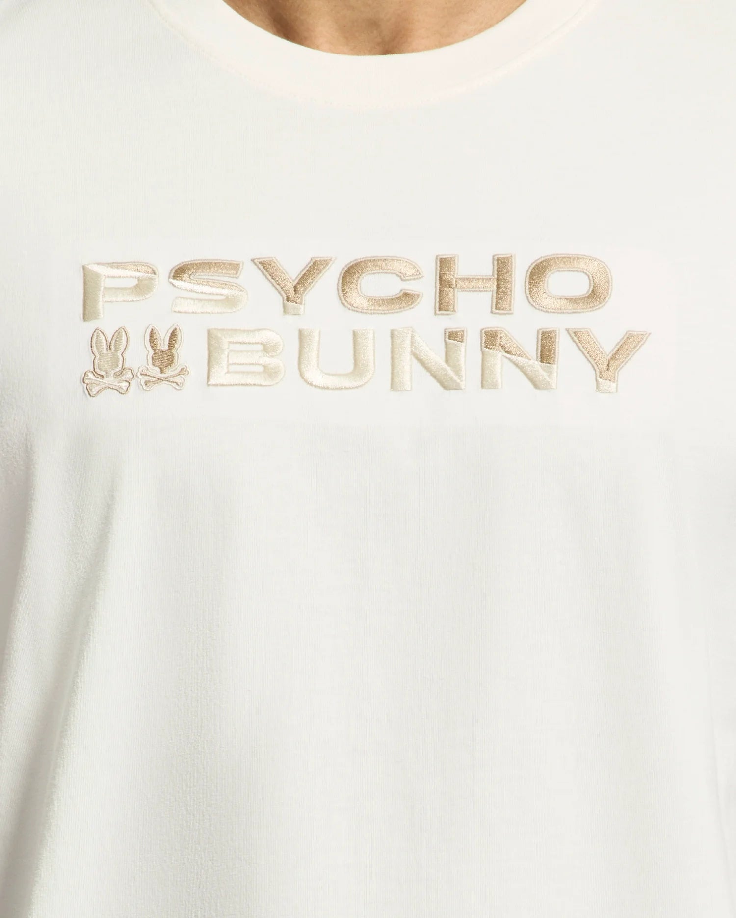Close-up of the Psycho Bunny Men's Berni Embroidered Wordmark Graphic Tee, crafted from soft Pima cotton. The shirt features "PSYCHO BUNNY" in beige stylized text and two small embroidered bunny head logos for a playful yet elegant touch.