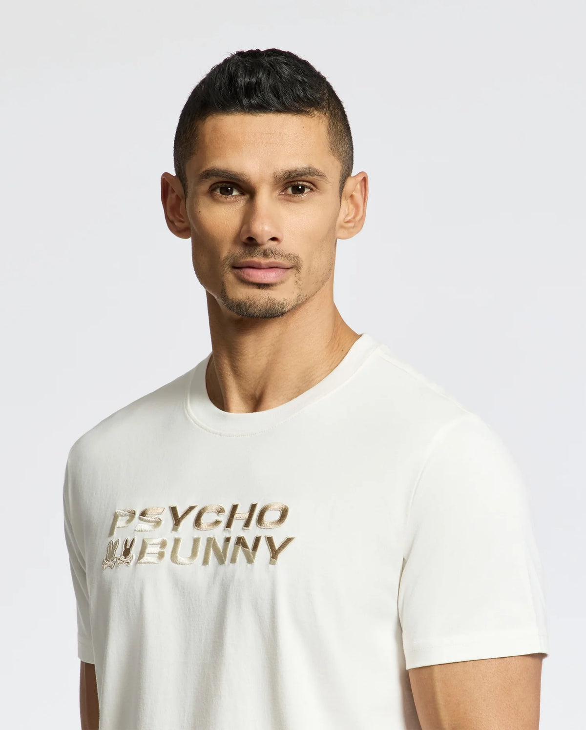 A man with short dark hair wears a white Psycho Bunny MENS BERNI EMBROIDERED WORDMARK GRAPHIC TEE crafted from Pima cotton, featuring "Psycho Bunny" in gold. An embroidered logo adds style as he stands against a plain white background, looking directly at the camera.