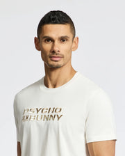 A man with short dark hair wears a white Psycho Bunny MENS BERNI EMBROIDERED WORDMARK GRAPHIC TEE crafted from Pima cotton, featuring "Psycho Bunny" in gold. An embroidered logo adds style as he stands against a plain white background, looking directly at the camera.