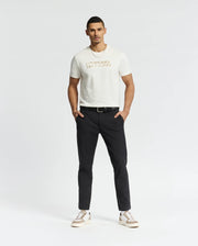 A man stands confidently against a plain background, wearing a white Psycho Bunny MENS BERNI EMBROIDERED WORDMARK GRAPHIC TEE made from Pima cotton with an embroidered logo. He pairs it with black pants, a belt, and white sneakers featuring beige accents, casually resting his hands in his pockets.