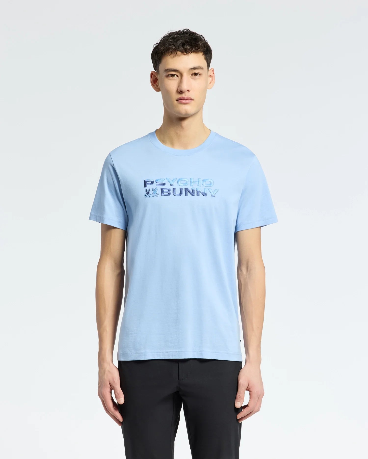 A young man wears a Psycho Bunny light blue Pima cotton tee, 'MENS BERNI EMBROIDERED WORDMARK GRAPHIC TEE - B6U417F200,' elegantly embroidered in dark blue, with black pants. The breathable fabric provides supreme comfort against a plain white backdrop.