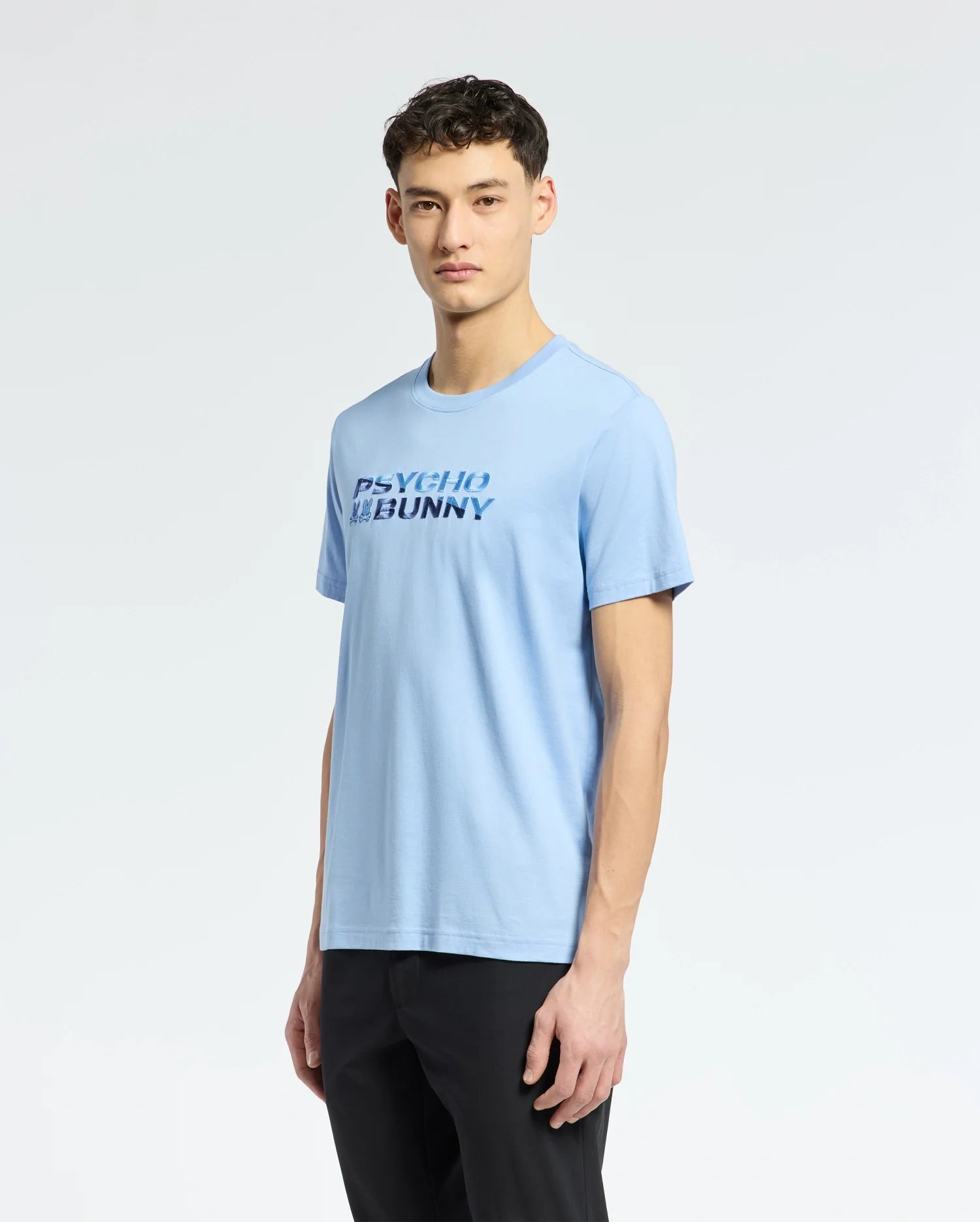 A man stands against a plain white background, wearing a light blue MENS BERNI EMBROIDERED WORDMARK GRAPHIC TEE (B6U417F200) by Psycho Bunny.