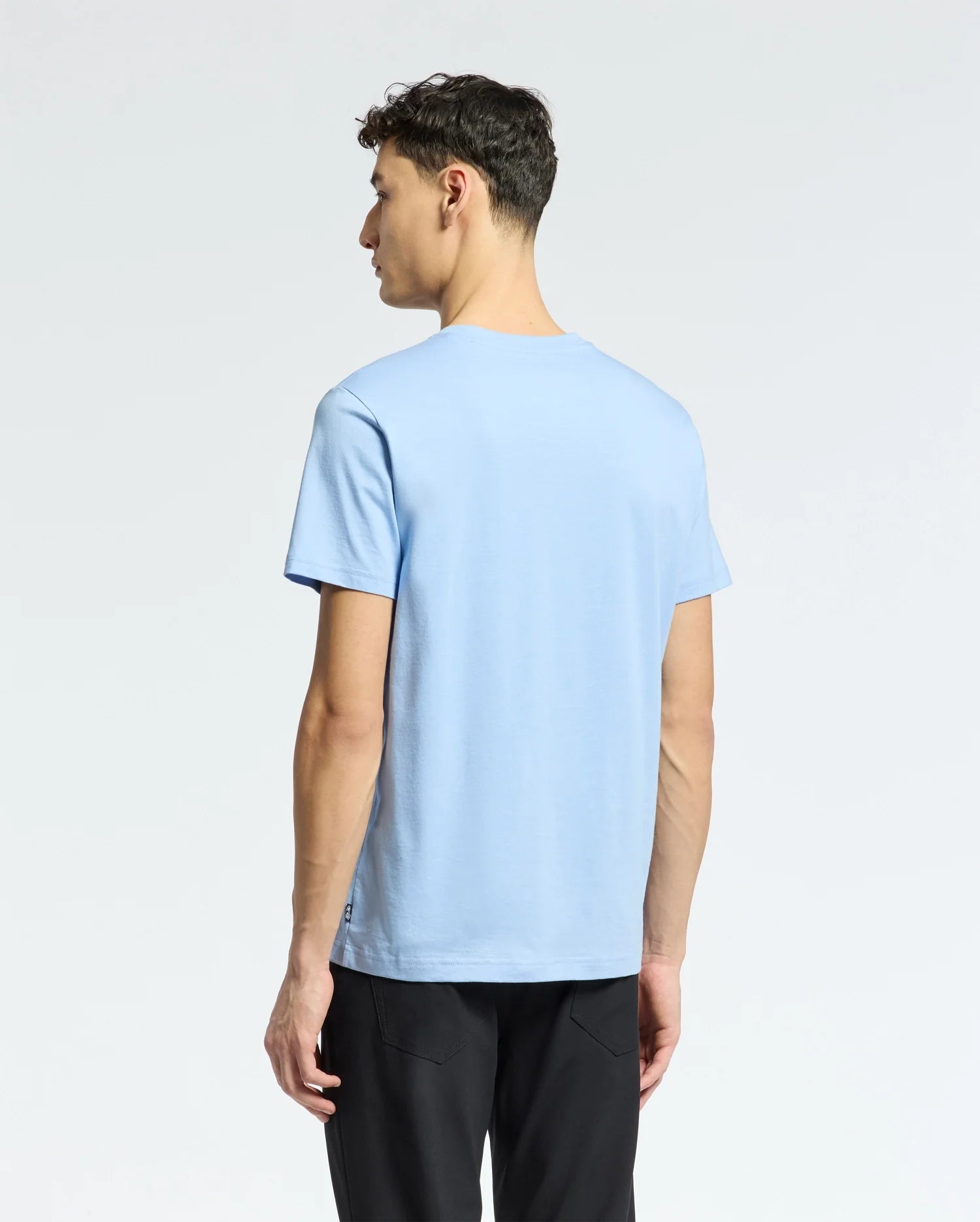 A person with short, dark hair faces away in a light blue Psycho Bunny MENS BERNI EMBROIDERED WORDMARK GRAPHIC TEE and black pants against a plain, light gray background.
