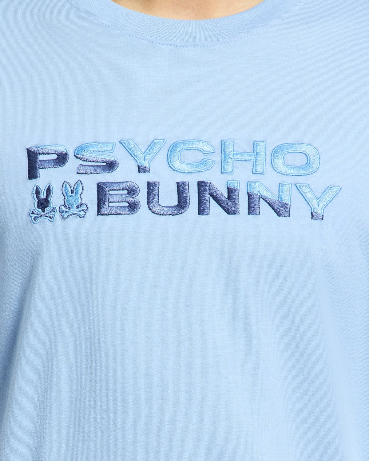 Close-up of the breathable light blue Psycho Bunny Men's Berni Embroidered Wordmark Graphic Tee featuring the embroidered text "PSYCHO BUNNY" in blue and two stylized bunny logos between the words.