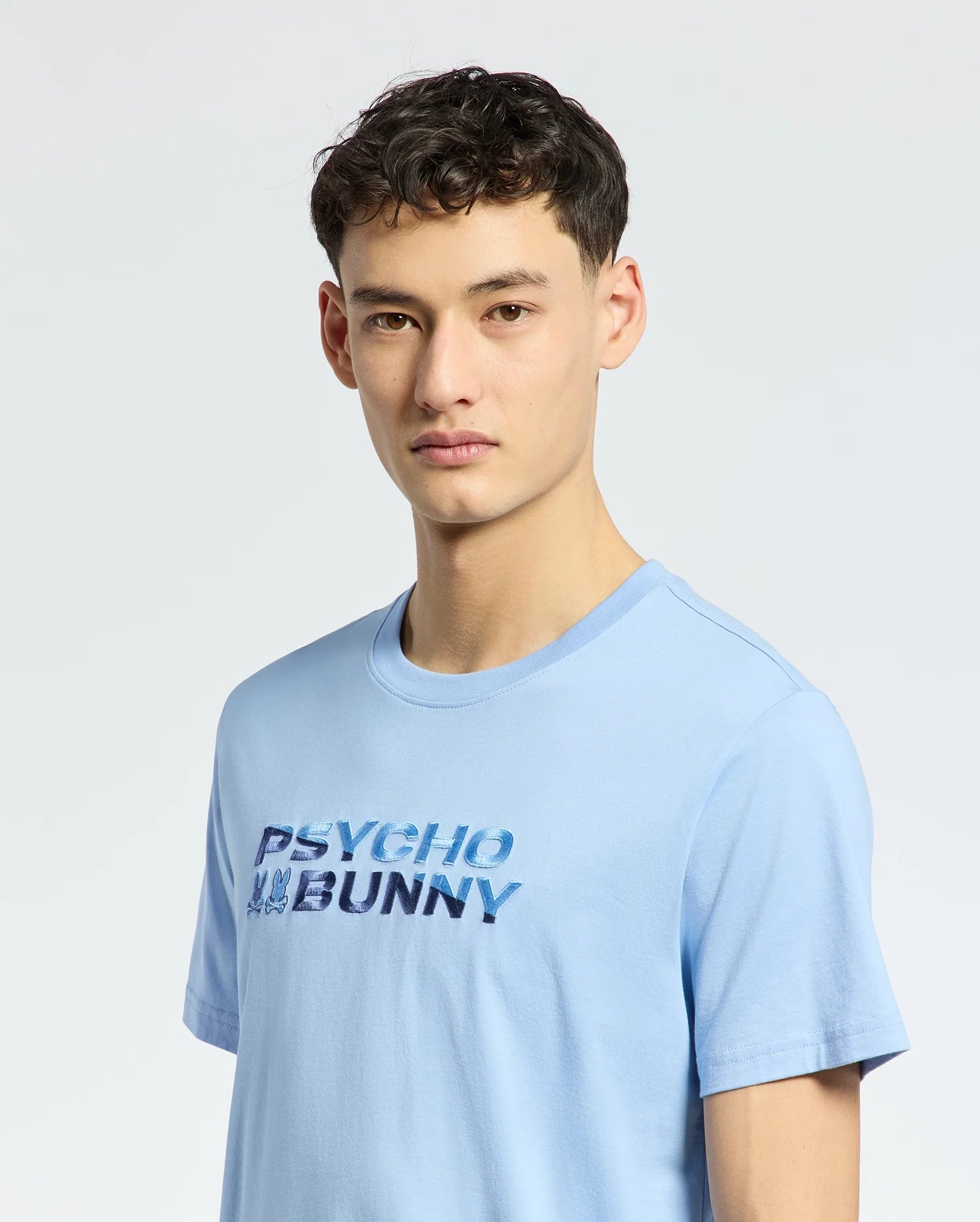 A young person with short, dark hair wears a light blue Psycho Bunny Mens Berni Embroidered Wordmark Graphic Tee, B6U417F200. The breathable Pima cotton ensures comfort against a plain light gray background.