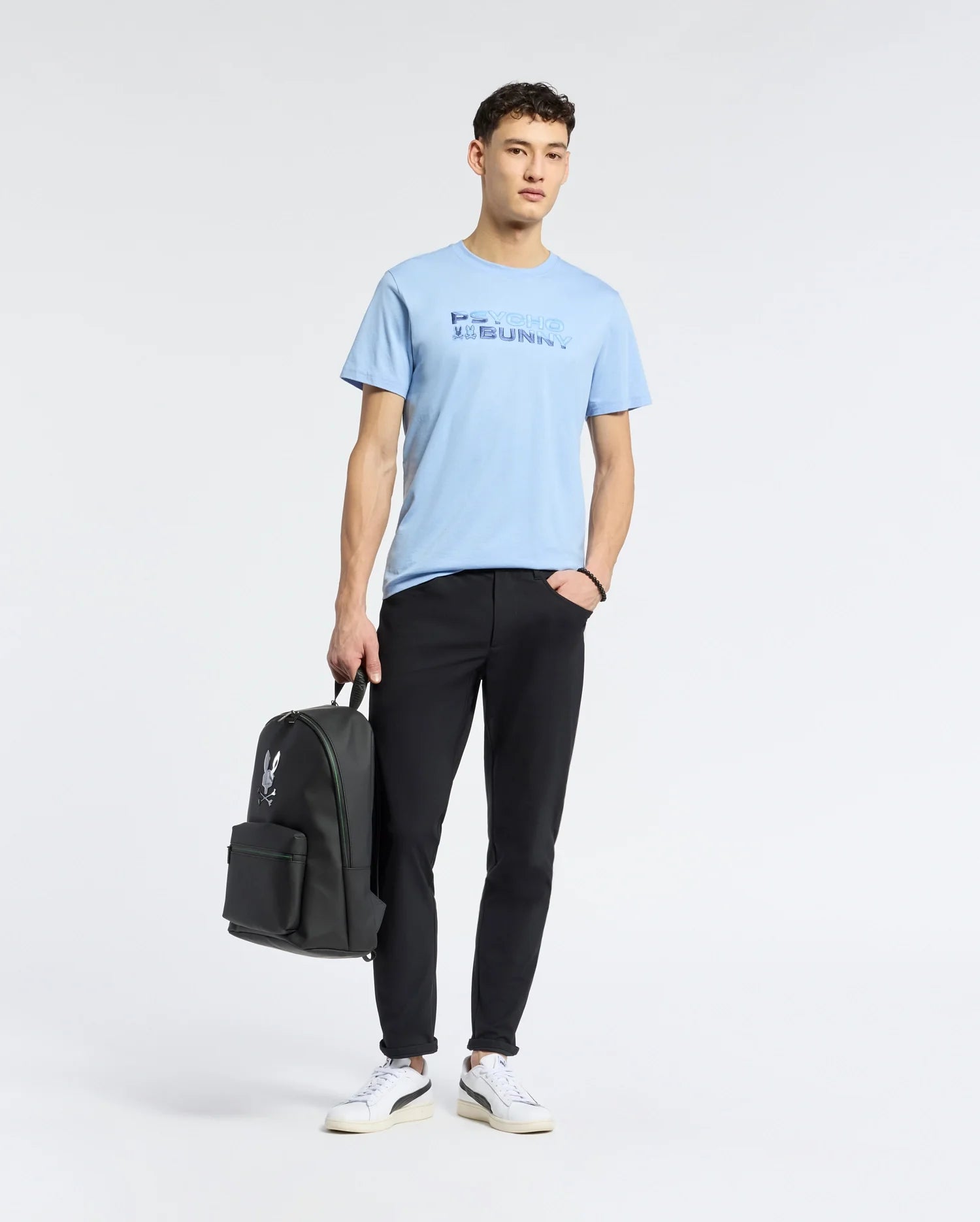 A young man in a breathable Psycho Bunny Men's Berni Embroidered Wordmark Graphic Tee, light blue Pima cotton, and black pants stands holding a black backpack. He wears white sneakers and gazes forward against a plain white background.