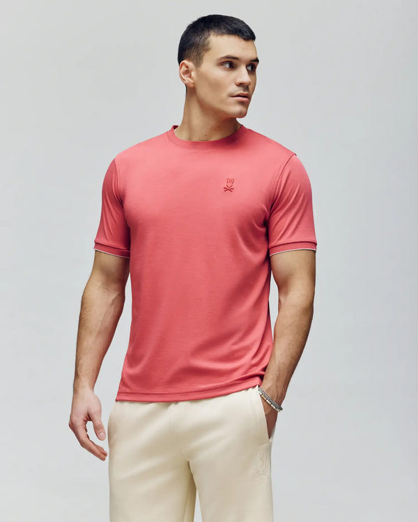 A man with short dark hair wears the Psycho Bunny MENS BRYCE PERFORMANCE TEE - B6U490F200 in red and cream pants, standing against a light gray background, looking left with his right hand in his pocket.
