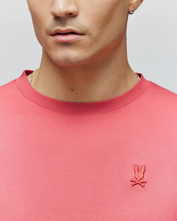 A close-up of a person wearing the Psycho Bunny MENS BRYCE PERFORMANCE TEE (B6U490F200) in pink, made from ultra-soft Pima cotton with an embroidered bunny skull and crossbones logo on the chest. The person has light skin, a silver hoop earring, and slight stubble.