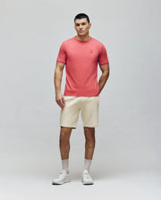 A person stands against a plain light background, wearing Psycho Bunny's MENS BRYCE PERFORMANCE TEE (B6U490F200) in pink, with light beige shorts, gray socks, and white sneakers—one hand in their pocket and a neutral expression.